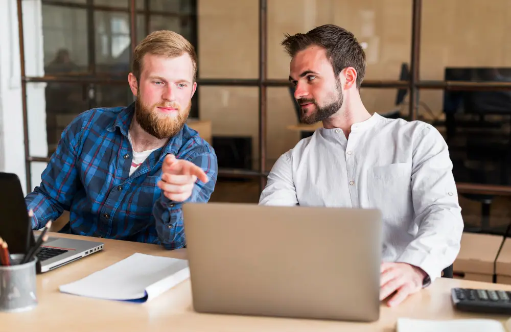 Two colleagues working at a laptop in an office, discussing strategy, planning, and digital project ideas.